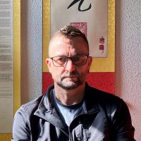 Person with short hair and glasses wearing a dark zip-up jacket over a light blue shirt, seated in front of a wall with calligraphy artwork bordered in red and gold.