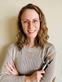 2. Person with wavy light brown hair and glasses smiles while holding several paintbrushes, wearing a beige knit sweater against a plain background.