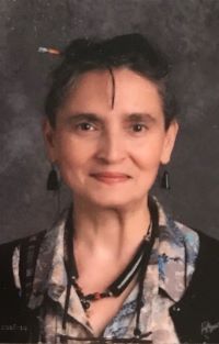 Person with dark hair pulled back smiles softly, wearing a patterned blouse, black earrings, and layered necklaces against a gray studio background.