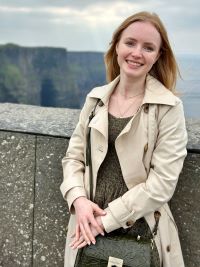 Person with light-colored hair smiling, wearing a beige trench coat over a green dress and holding a floral handbag. Misty cliffs and stone barrier visible in the background.