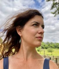 Person with long, dark hair blowing in the wind, wearing a sleeveless top and teardrop-shaped earring, standing outdoors in front of a green field and trees. The sky is partly cloudy with patches of blue.