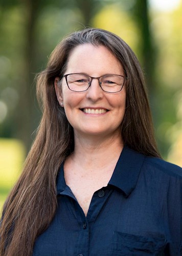 Professional headshot of a person with long brown hair, glasses, and a navy-blue top, posed against a green background of natural lighting.