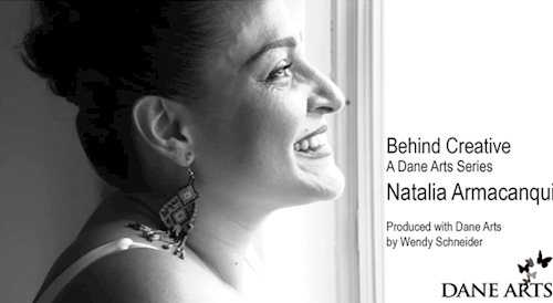 Black and white image of a smiling woman with dark hair in a bun and beaded earrings looking out a brightly lit window