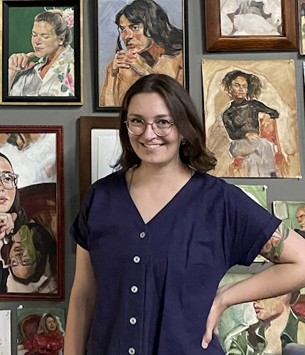 A person wearing glasses and a dark blue buttoned shirt stands smiling with one hand on their hip in front of a wall filled with expressive portrait paintings. The artworks vary in size and framing, depicting different individuals in warm and cool tones, 