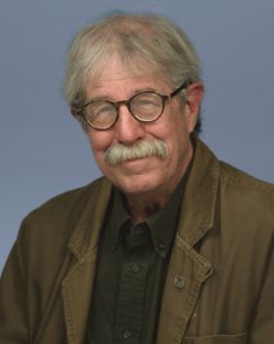 Professional headshot portrait of a man with short grey hair, black circular glasses, and a white mustache wearing a brown jacket. 
