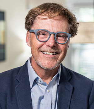 Headshot of a smiling man with reddish-blonde hair wearing glasses and a dark blue suit