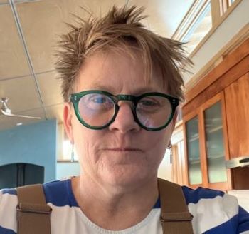 Close-up headshot of a person with short, spiky light brown hair and round dark-framed glasses, wearing a white top with blue stripes and tan overall straps, indoors in front of warm wood cabinetry and a blue wall.