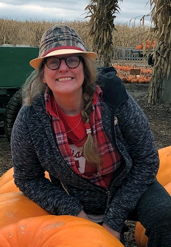 Photo of a woman with a long brown braid wearing glasses, a hat, and a red shirt with grey sweatshirt. She is surrounded by pumpkins and large stalks of corn with a tractor in the background.